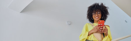 Pretty African-American woman in yellow sweater holds phone leaning onto railing on staircaseの写真素材