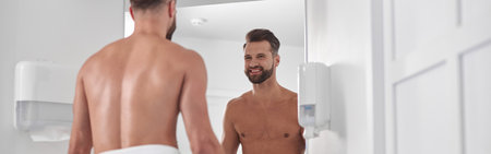 Happy bearded man with bare chest wrapped with towel looks in mirror in modern barthroomの写真素材