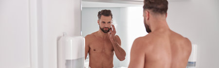 Attractive middle aged man applies cream onto face looking in mirror in bathroomの写真素材