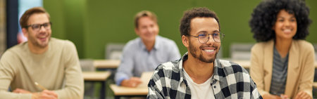 Young cheerful mixed race male office worker sitting listening to coach during seminar or training and smiling while studying with colleagues in coworking spaceの写真素材