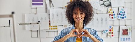 African-American woman designer shows heart with hands near sewing machine in workshopの写真素材