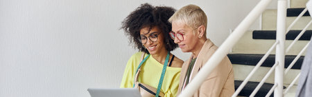 Mature woman and young African-American use laptop stitting on staircase in design studioの写真素材