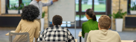 Confident mature coach or speaker pointing at flip chart and explaining something to people sitting in front of him in coworking space during corporate training or seminarの写真素材