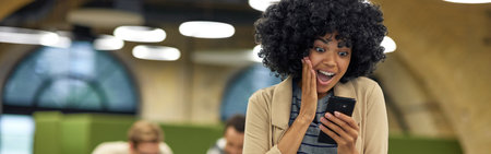 Good news. Young happy mixed race woman looking at smartphone and feeling excited while standing in coworking spaceの写真素材