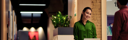 Young cheerful asian woman talking with colleague while standing together in corridor of the modern officeの写真素材