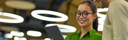 Young cheerful asian woman showing something on digital tablet to her male colleague while standing together in the coworking space or officeの写真素材