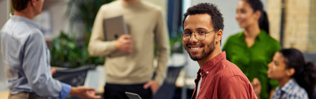 Young cheerful mixed race man holding digital tablet and smiling at camera while working with colleagues in the modern office, selective focusの写真素材