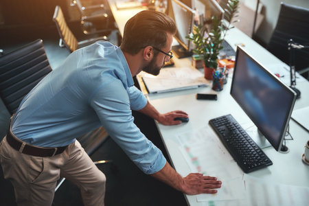 Checking e-mail. Young bearded businessman in eyeglasses and formal wear using computer while standing in the modern officeの写真素材