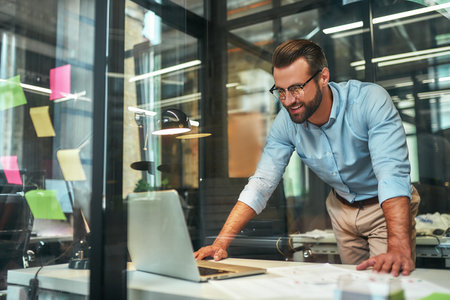 Great results Young bearded businessman in eyeglasses and formal wear looking at laptop and smiling while standing in the modern officeの写真素材