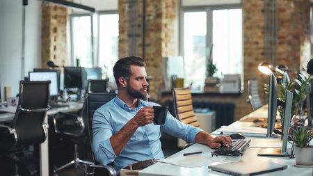 Fresh coffee. Side view of young bearded businessman in formal wear holding cup of coffee and looking at computer while sitting in the modern officeの写真素材
