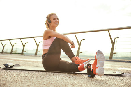 Happy morning. Positive disabled athlete woman with prosthetic leg in headphones doing yoga exercises and smiling while sitting on the bridgeの写真素材