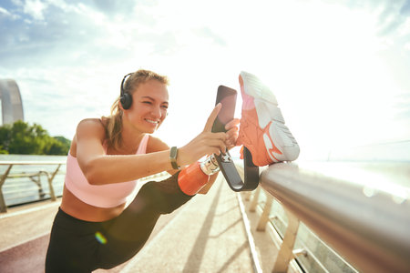 Staying in touch. Happy disabled woman in sportswear and headphones stretching prosthetic leg and looking with smile at her smartphone while standing on the bridgeの写真素材