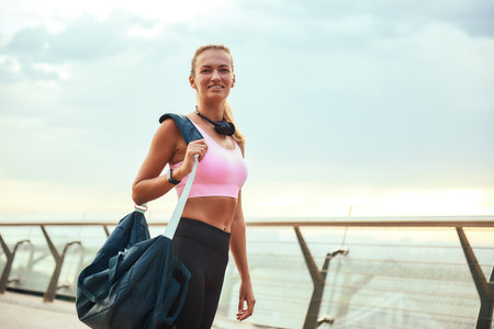 Full of energy. Beautiful young woman in sports clothing carrying her sport bag and smiling while standing on the bridge in the morningの写真素材