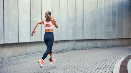 On the way to success.Back view of young disabled woman with leg prosthesis in comfortable sports clothing is running outdoors along the streetの写真素材