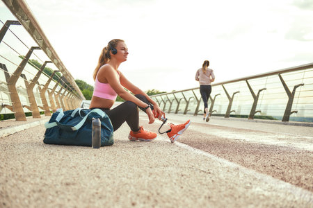 Time to relax. Side view of young beautiful woman in headphones with leg prosthesis listening music and looking aside with smile while sitting on the bridgeの写真素材