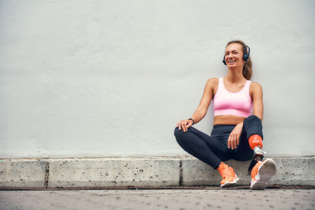 Simply happy Young smiling woman with leg prosthesis in sports clothing and headphones listening music after her workout while sitting outdoorsの写真素材