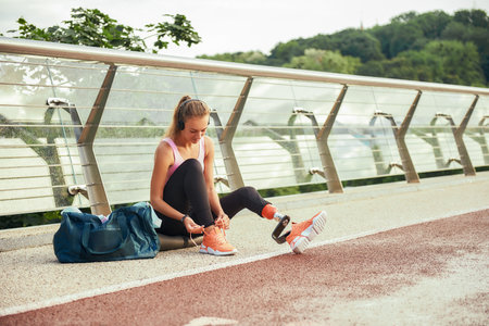 Getting ready to run. Young woman in sports clothing and headphones with leg prosthesis tying her shoelaces while sitting on the bridgeの写真素材
