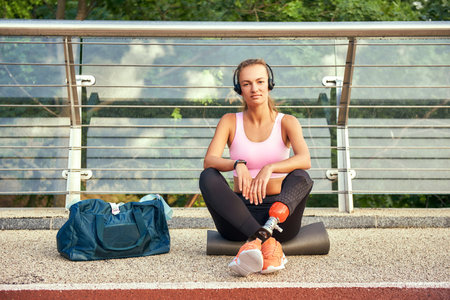Confident in myself. Portrait of young woman in sports clothing and headphones with leg prosthesis sitting on the bridge and looking at cameraの写真素材