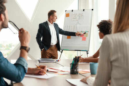 Analyzing results. Mature businessman standing near whiteboard and explaining something to his colleaguesの写真素材
