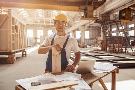 Cheerful builder standing by the table with laptop and papers