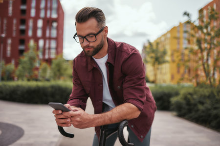 I am always in touch. Handsome man with stubble in casual clothes and eyeglasses leaning at his bicycle and looking at mobile phone while standing outdoorsの写真素材