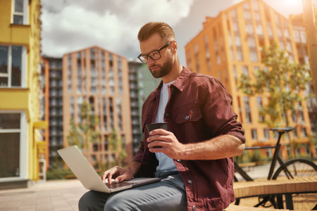 I need coffee. Handsome man with stubble in casual clothes and eyeglasses working on laptop and holding cup of coffee while sitting on the bench outdoorsの写真素材