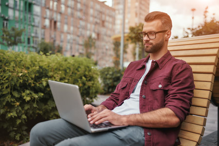 Modern blogger. Side view of young handsome man with stubble in casual clothes and eyeglasses working on laptop while sitting on the bench outdoorsの写真素材