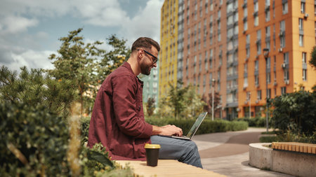 Happy and young. Side view of handsome man with stubble in casual clothes and eyeglasses working on laptop while sitting on the bench outdoorsの写真素材