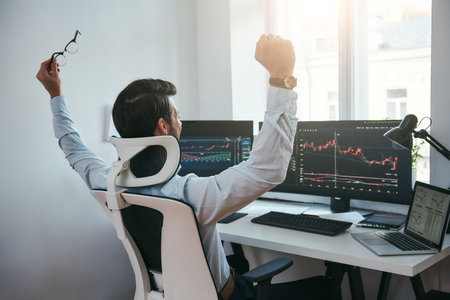 Yes Back view of stock trader with raised hands looking at multiple computer screens with data and charts and feeling happy while sitting in modern officeの写真素材