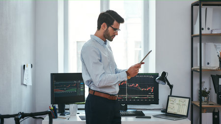 Analyzing data. Successful young trader in formal wear is looking at trading graphs and reports on his digital tablet while standing in front of computer screens with trading charts in the office.の写真素材