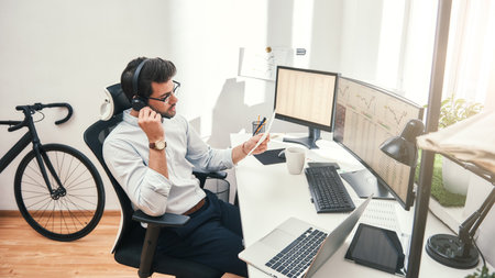 Talking about business. Side view of young bearded trader in headset holding financial report and talking with client while sitting in front of monitor screens in the officeの写真素材