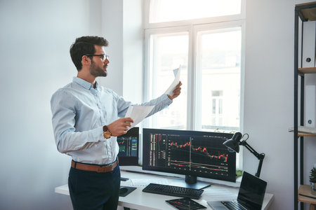 Trading strategy. Smart and young trader in eyeglasses looking at financial reports and analyzing trading charts while standing in front of computer screens in modern office.の写真素材