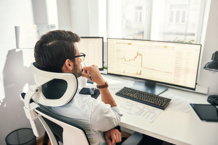 No time to relax. Back view of professional trader working with graph and financial reports on computer screens while sitting in his modern office.の写真素材