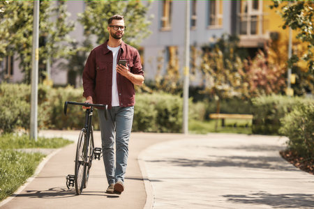 My way. Handsome man with stubble in casual clothes and eyeglasses pulling his bicycle and holding smartphone while walking outdoorsの写真素材