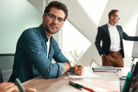 Young and handsome man in eyeglasses is looking at camera and smiling while his boss standing near whiteboard and explaining something on the backgroundの写真素材