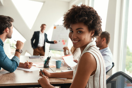 Young afro american woman is looking at camera and smiling while her boss standing near whiteboard and discussing something with teamの写真素材