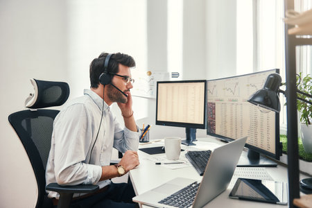 Always available. Side view of young bearded trader in headset talking with client and looking at...