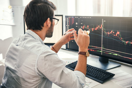 Successful trading. Back view of young bearded trader in formal wear pointing on the data on computer screen with pen and holding Bitcoin in one hand while working his modern office.の写真素材
