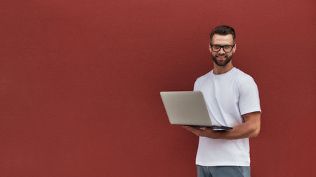 Happy blogger. Handsome young man in casual clothes and eyeglasses holding laptop and looking at camera with smile while standing against red wall outdoorsの写真素材