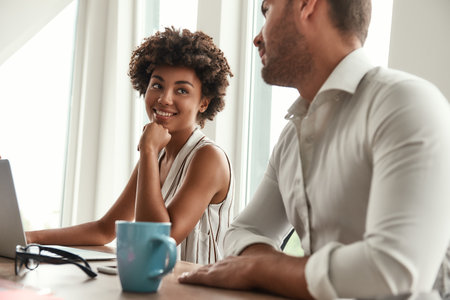 New working day. Young and beautiful afro american woman discussing something with her colleague while sitting in the modern officeの写真素材