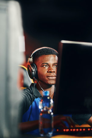Fully concentrated young african guy, professional cybersport gamer wearing headphones sitting in gaming club or internet cafe and playing online video gamesの写真素材