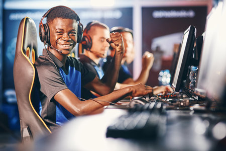 Side view of a cheerful african guy, professional cybersport gamer wearing headphones looking at camera and smiling while participating with team in eSport tournamentの写真素材