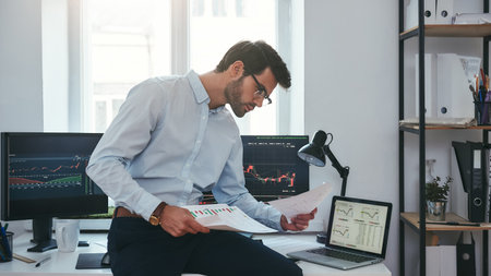 Busy working day. Young successful trader or businessman in formal wear and eyeglasses sitting at modern office in front of computer screens with charts and reading market reportの写真素材