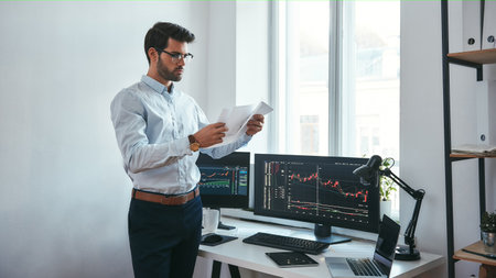 Analyzing data. Thoughtful trader in eyeglasses holding a financial report and analyzing trading charts while standing in front of computer screens in modern officeの写真素材