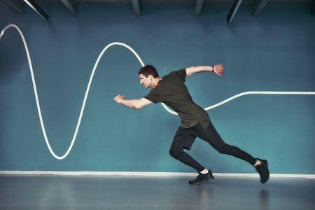 Great shape. Full length of young athletic man in sport clothing running against blue wall while...