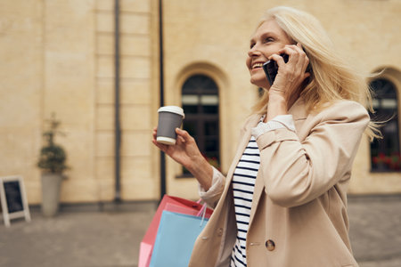 Mature woman walking in city street with coffee and talking phone after shopping. Seasonal saleの写真素材