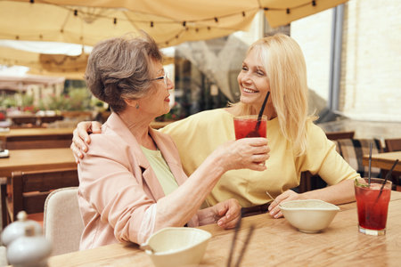 Smiling mature ladies sitting in cafe terrace talking and hugging while drinking cocktailsの写真素材