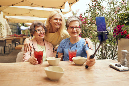 Mature female friends sitting in cafe terrace during lunch time and making selfie on phoneの写真素材