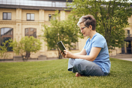 Senior woman in eyeglasses reading book while sitting on grass in park and having a restの写真素材