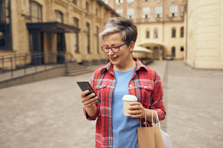 Stylish mature woman walking with her shopping bags, drinking coffee and use phone in city streetの写真素材